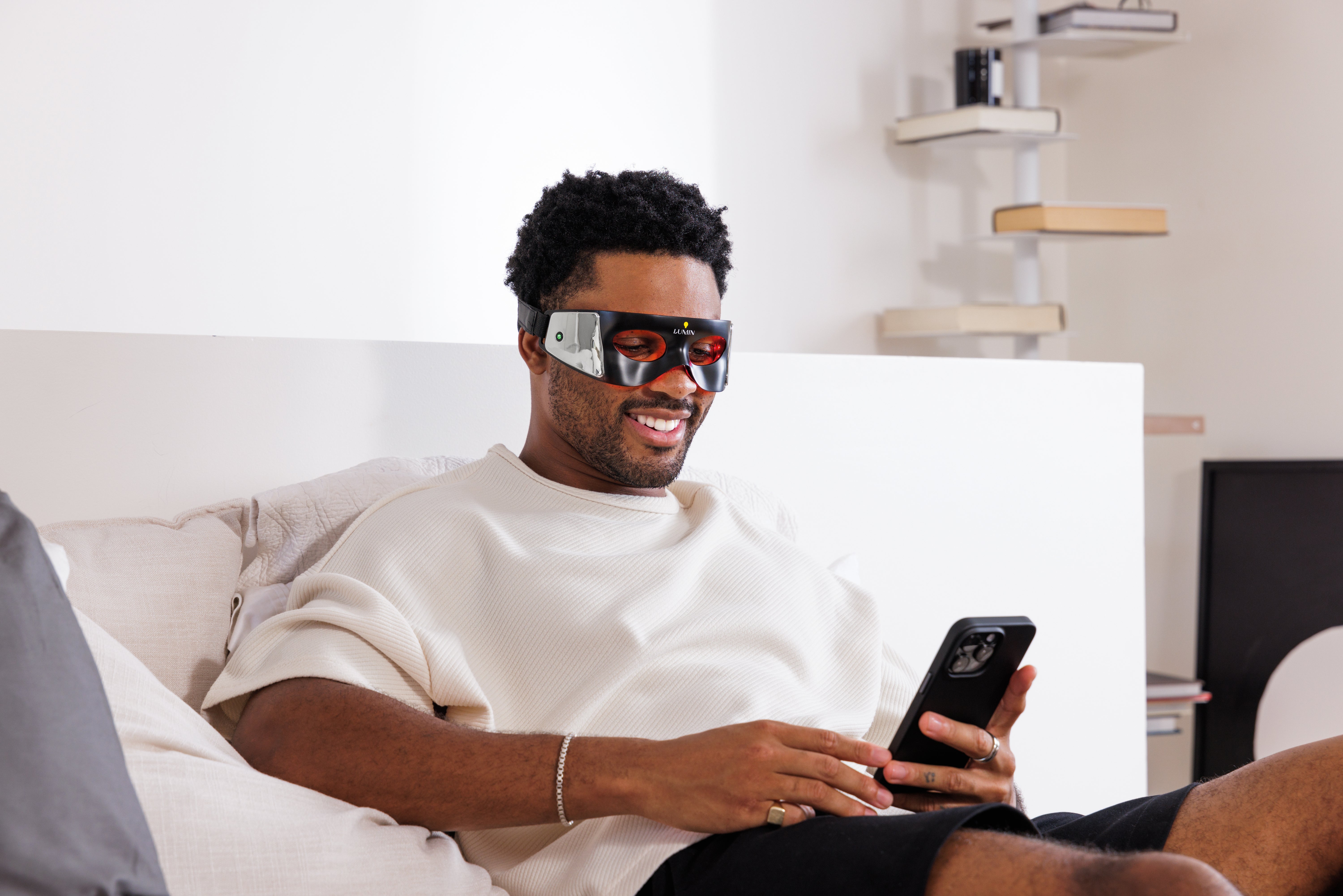 Man wearing futuristic smart glasses using smartphone while sitting on beige sofa in bright living room