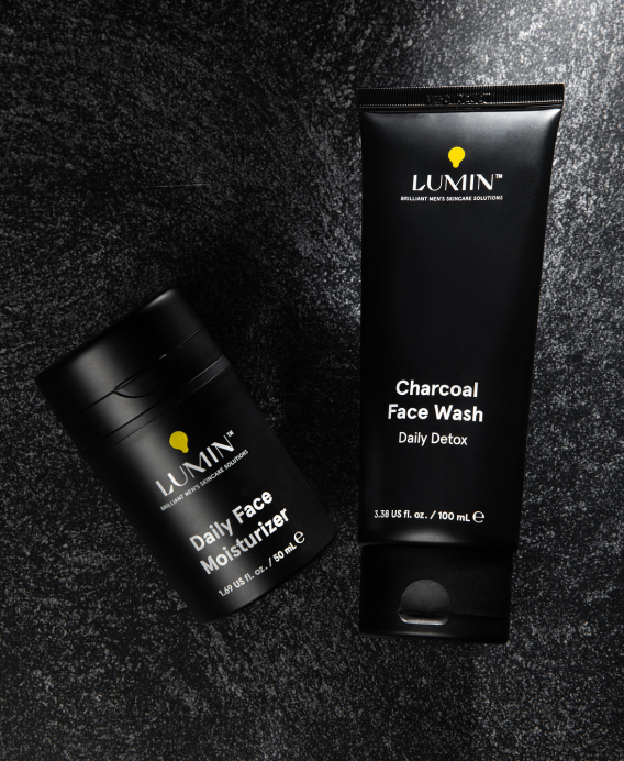 Charcoal face wash and daily face moisturizer with black packaging on black textured background for men's skincare.