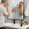 Middle-aged man applying facial skincare cream in modern bathroom with mirror, black faucet, and toiletry set on sink.