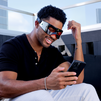 Man wearing futuristic black Lumin eye massager using smartphone outdoors with casual black shirt and white pants