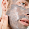 Man applying gray clay face mask on cheek with fingers, skincare routine, facial treatment for men, close-up.