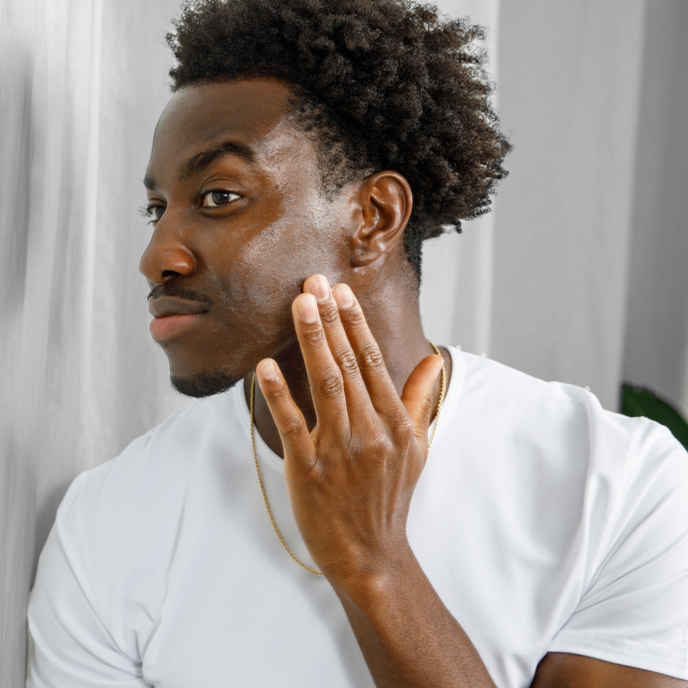 Young Black man applying moisturizer on face with hand, skincare routine, natural glow, white t-shirt, gold necklace