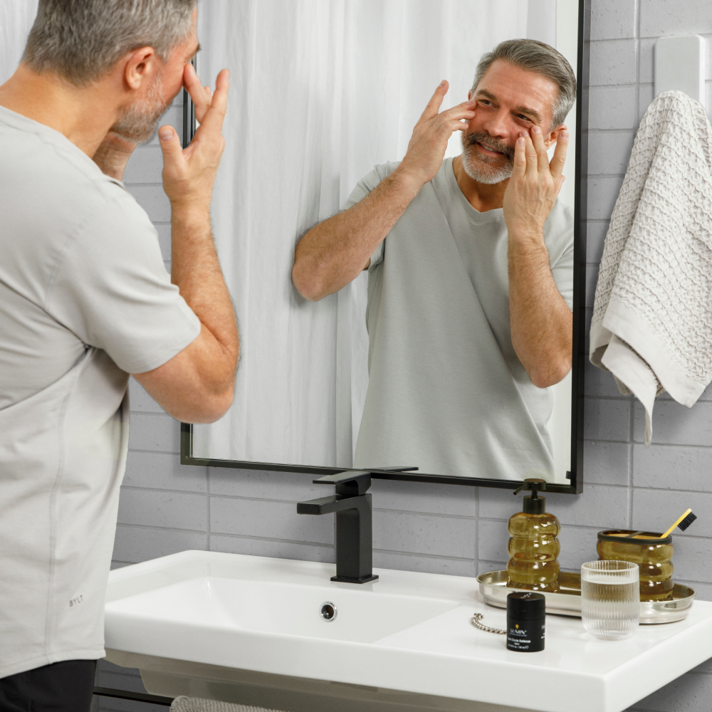 Middle-aged man applying anti-aging cream in bathroom mirror with skincare products on sink countertop.