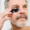 Mature man applying black under-eye patches for skin care, anti-aging, and dark circle treatment.