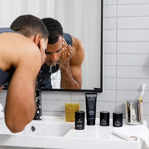 Man washing face in bathroom mirror with Lumin charcoal face wash, moisturizer, and skincare products on sink counter.