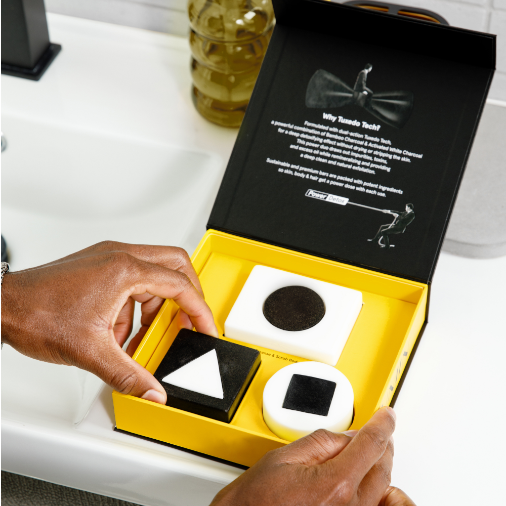 Black and white charcoal soap bars with geometric shapes in yellow detox skincare gift box, held by hands near bathroom sink.