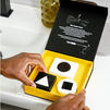 Black and white charcoal soap bars with geometric shapes in yellow detox skincare gift box, held by hands near bathroom sink.