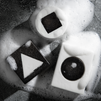 Black and white soap bars with geometric shapes—triangle, square, circle—surrounded by white foam on textured stone.