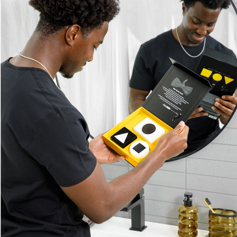Man holding open black and yellow grooming kit box with black and white geometric-shaped soaps near bathroom sink and mirror.