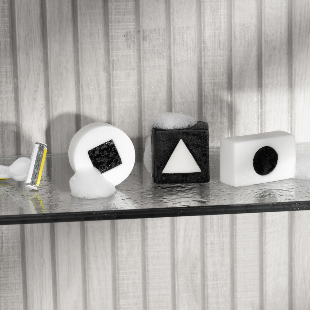 Razor and three black-and-white geometric soap bars with square, triangle, and circle shapes on glass shelf with foam bubbles.