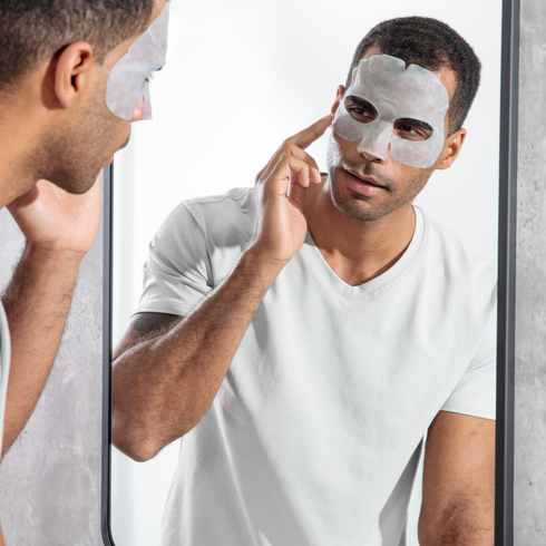 Man applying white sheet face mask under eyes and nose, wearing white t-shirt, skincare routine in bathroom mirror.