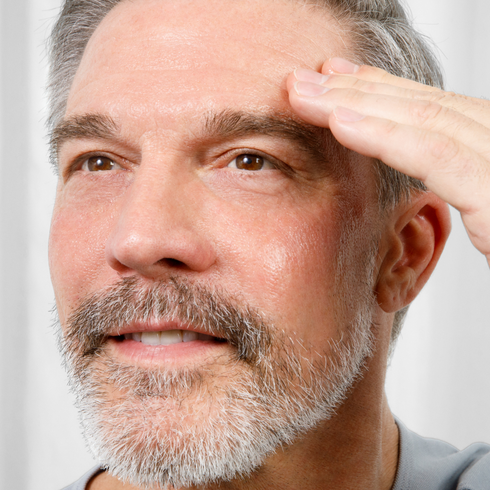 Mature man with gray beard gently touching forehead, showcasing skincare or anti-aging facial treatment for men.