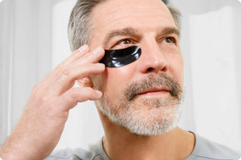 Mature man applying black under-eye mask patch for skincare, reducing puffiness and dark circles.