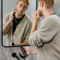 Young man grooming facial hair using electric trimmer in front of bathroom mirror, beige sweater, tattooed arm visible